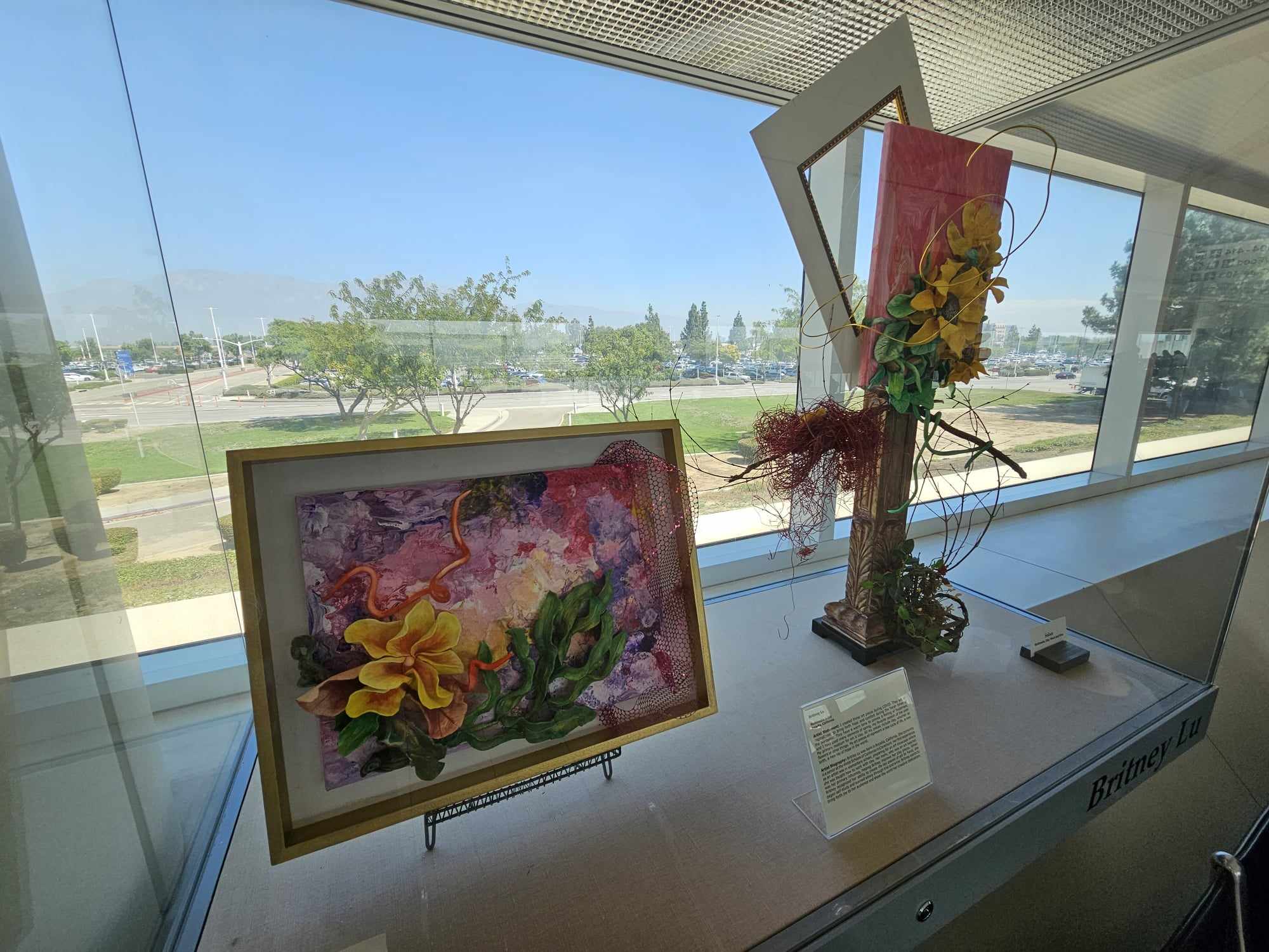 Framed floral painting and potted plant displayed in a bright room with outdoor view.