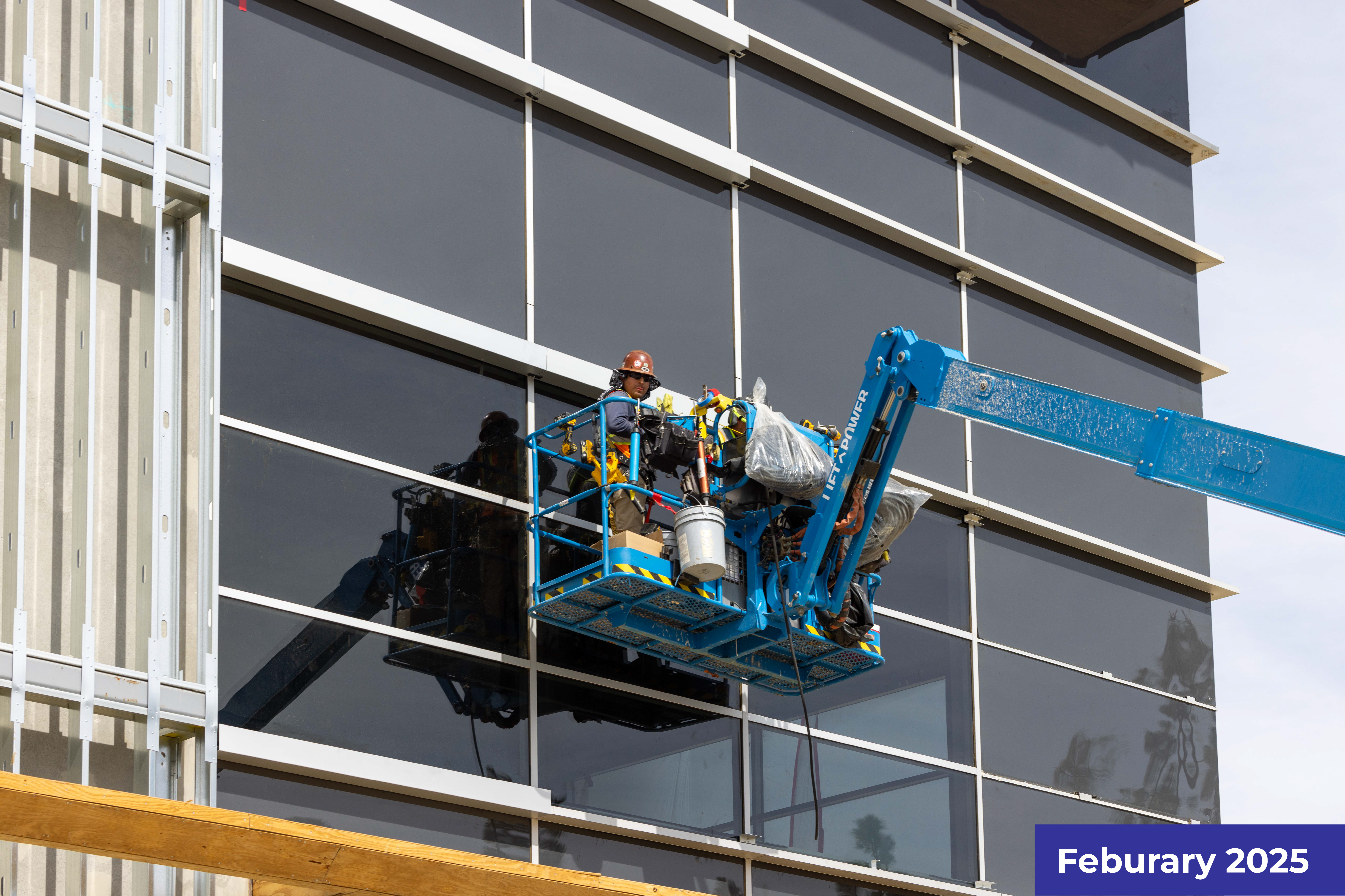 Construction workers on blue aerial lift installing glass panels on a building facade.