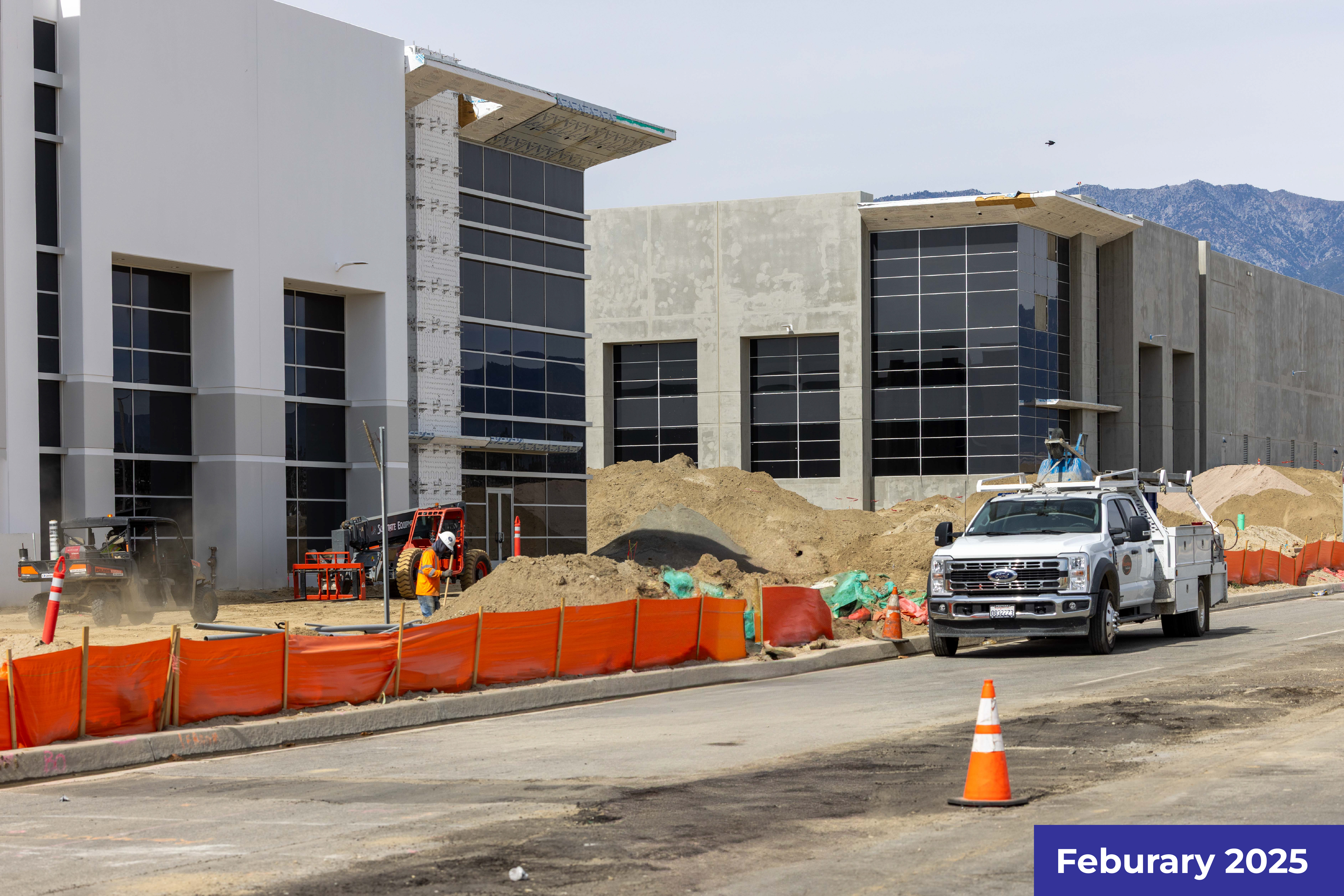 Modern commercial buildings under construction with orange safety barriers and construction equipment visible.