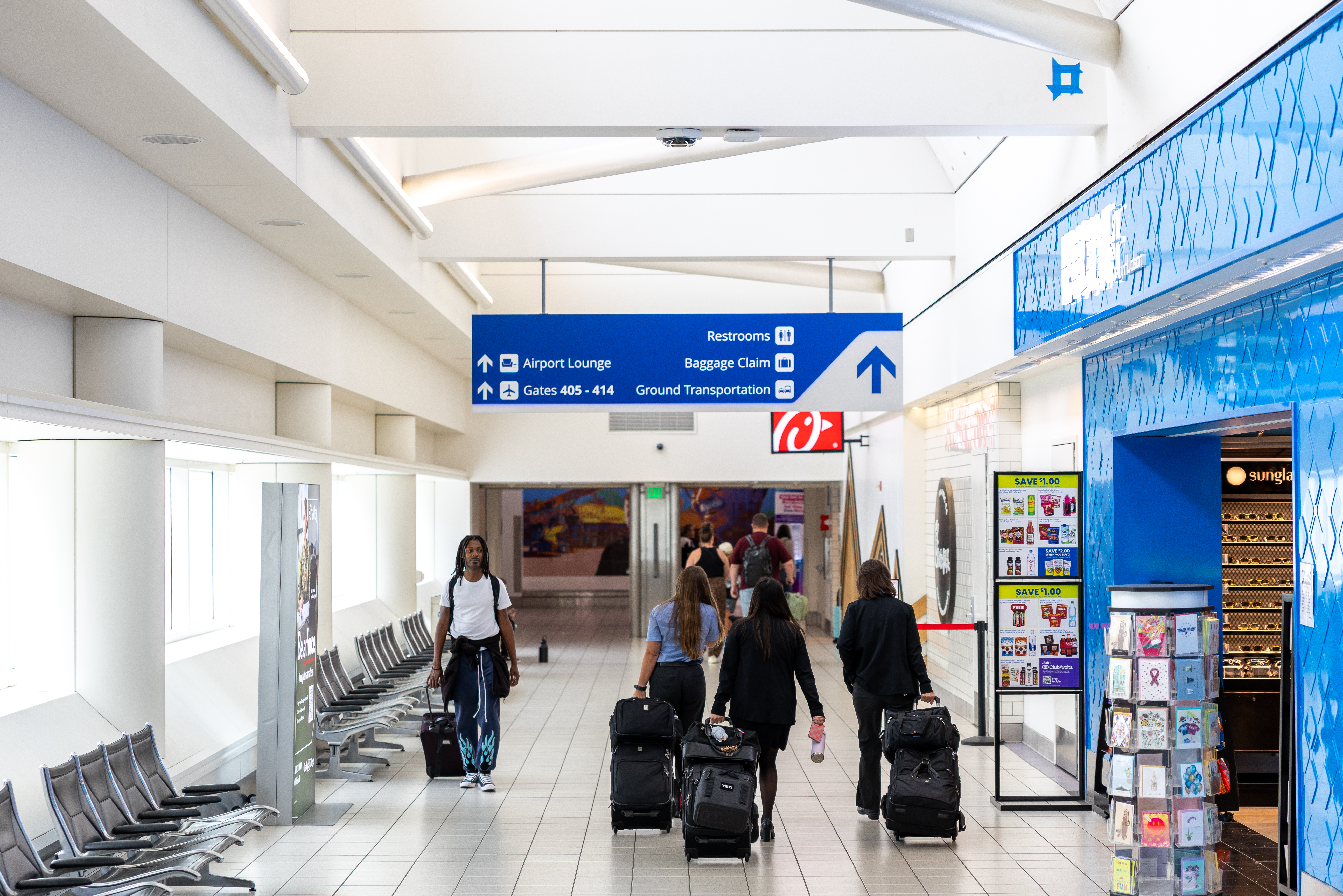 Airport terminal with travelers walking through a hallway with blue directional signs overhead.