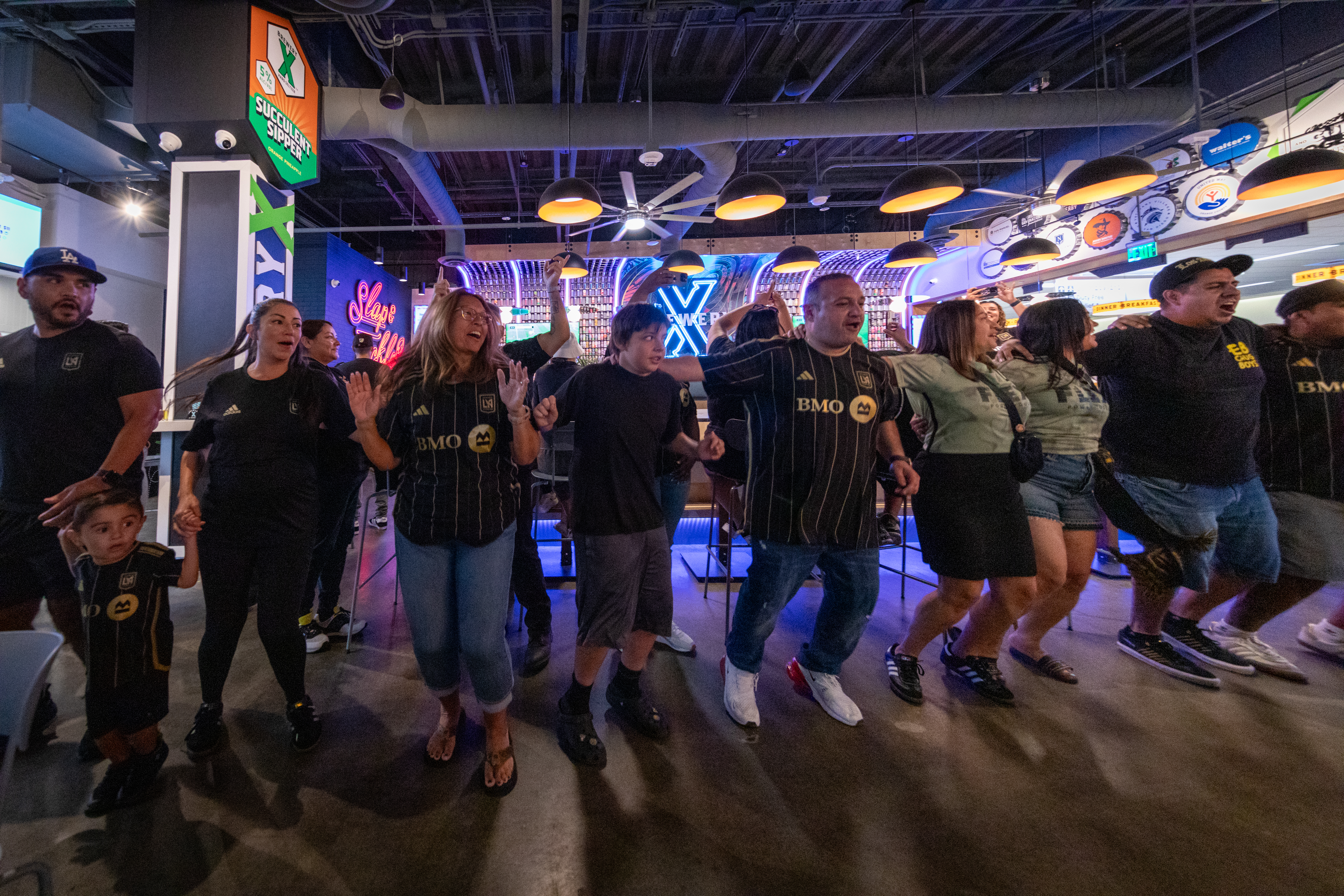 A line of people with arms over each other's shoulders dance in celebration of LAFC inside Brewery X at ONT.