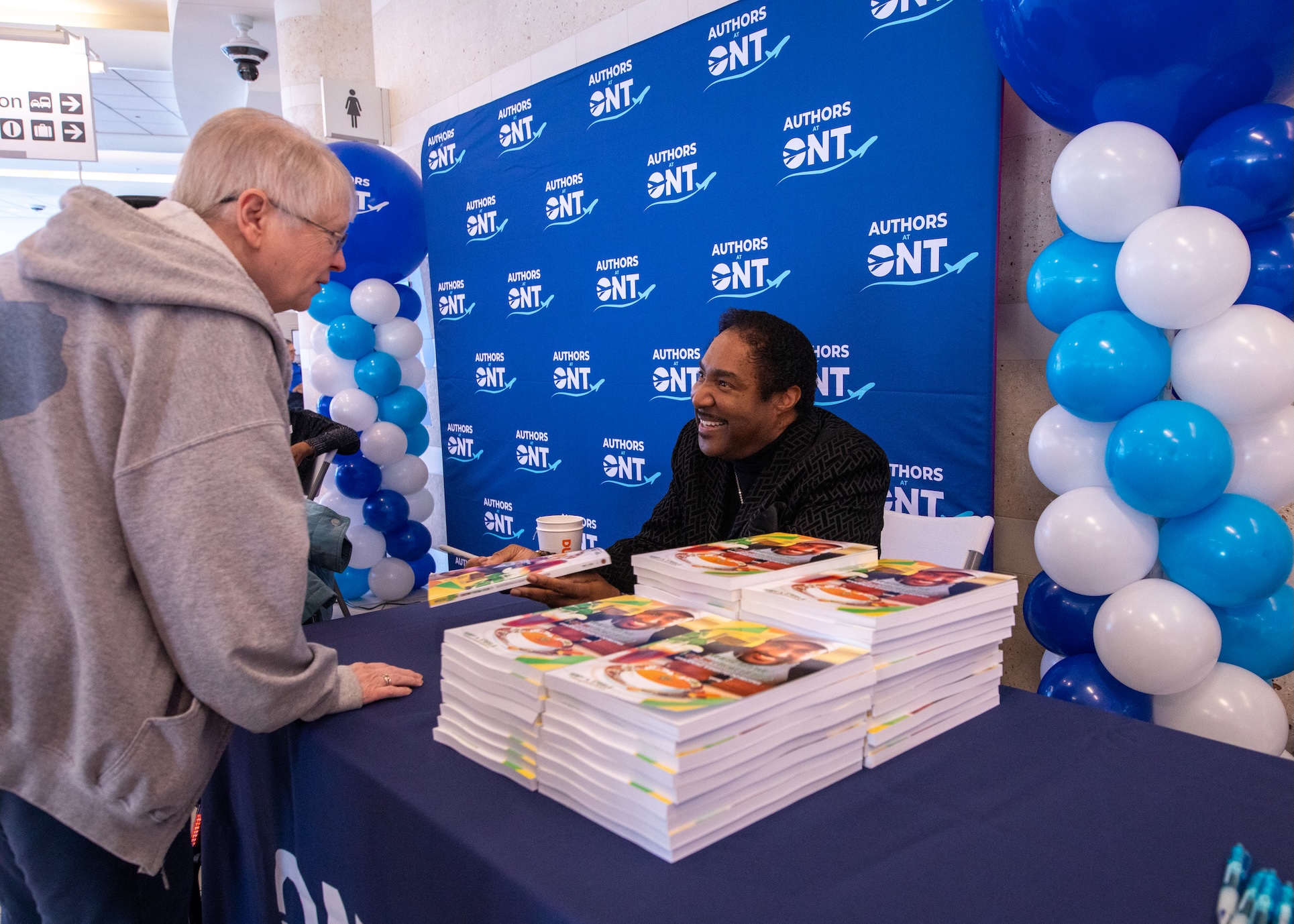 Grammy nominee Terry Steele smiles as he greets a passenger at a table filled with his books at a booth inside an ONT terminal.