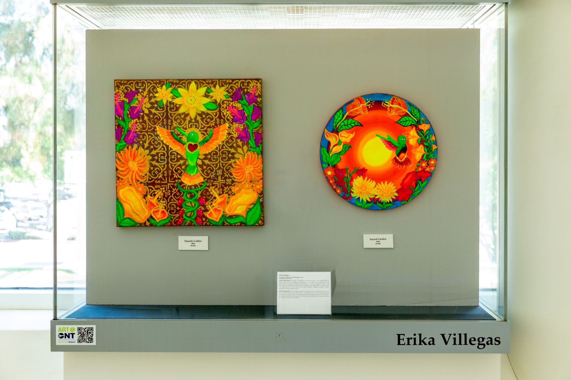 A brightly colored painting and object hang in a display case with artist Erika Villegas' name.
