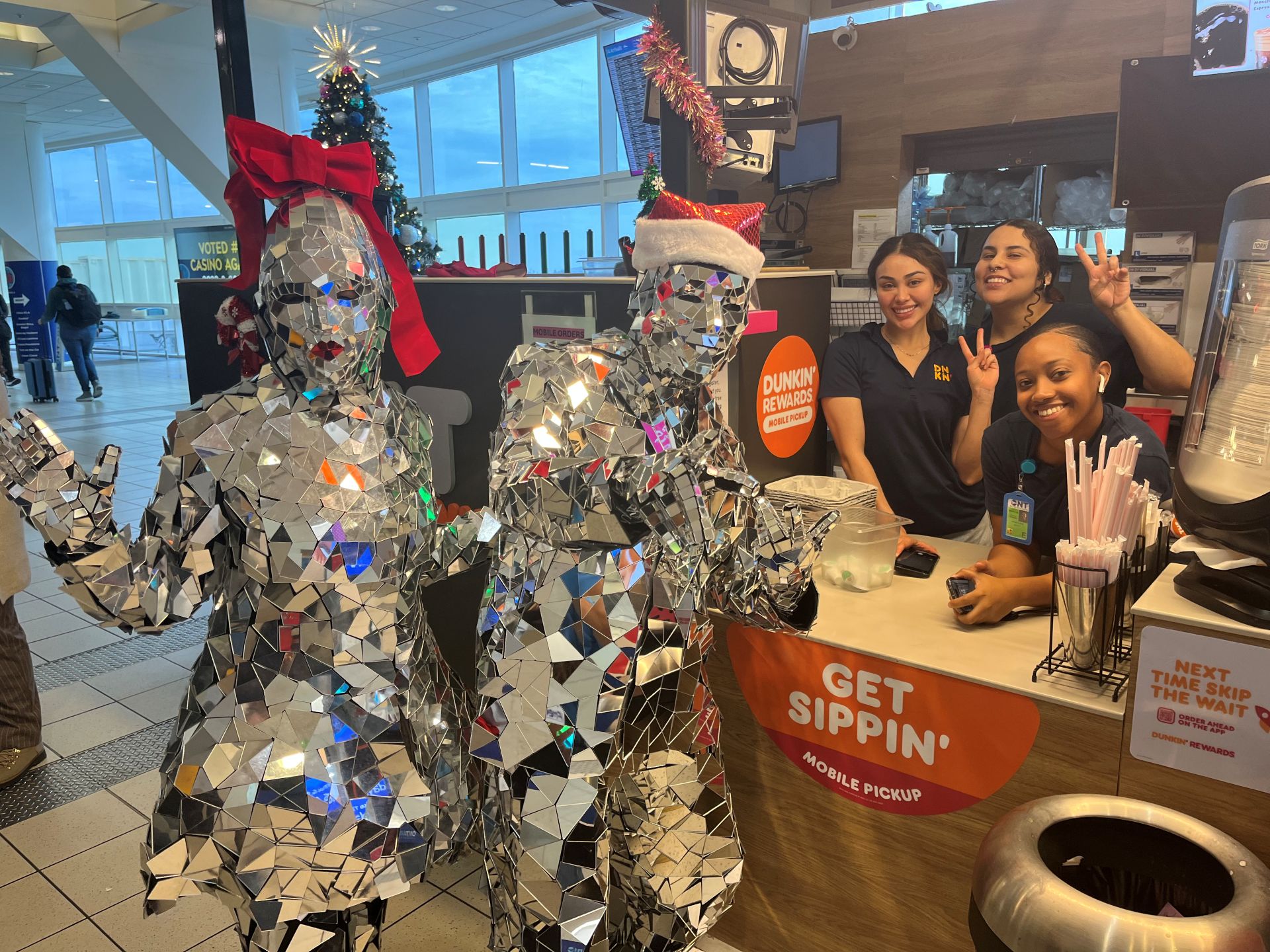 A man and a woman covered in sparkly silver costumes and wearing santa hats pose with Dunkin workers.