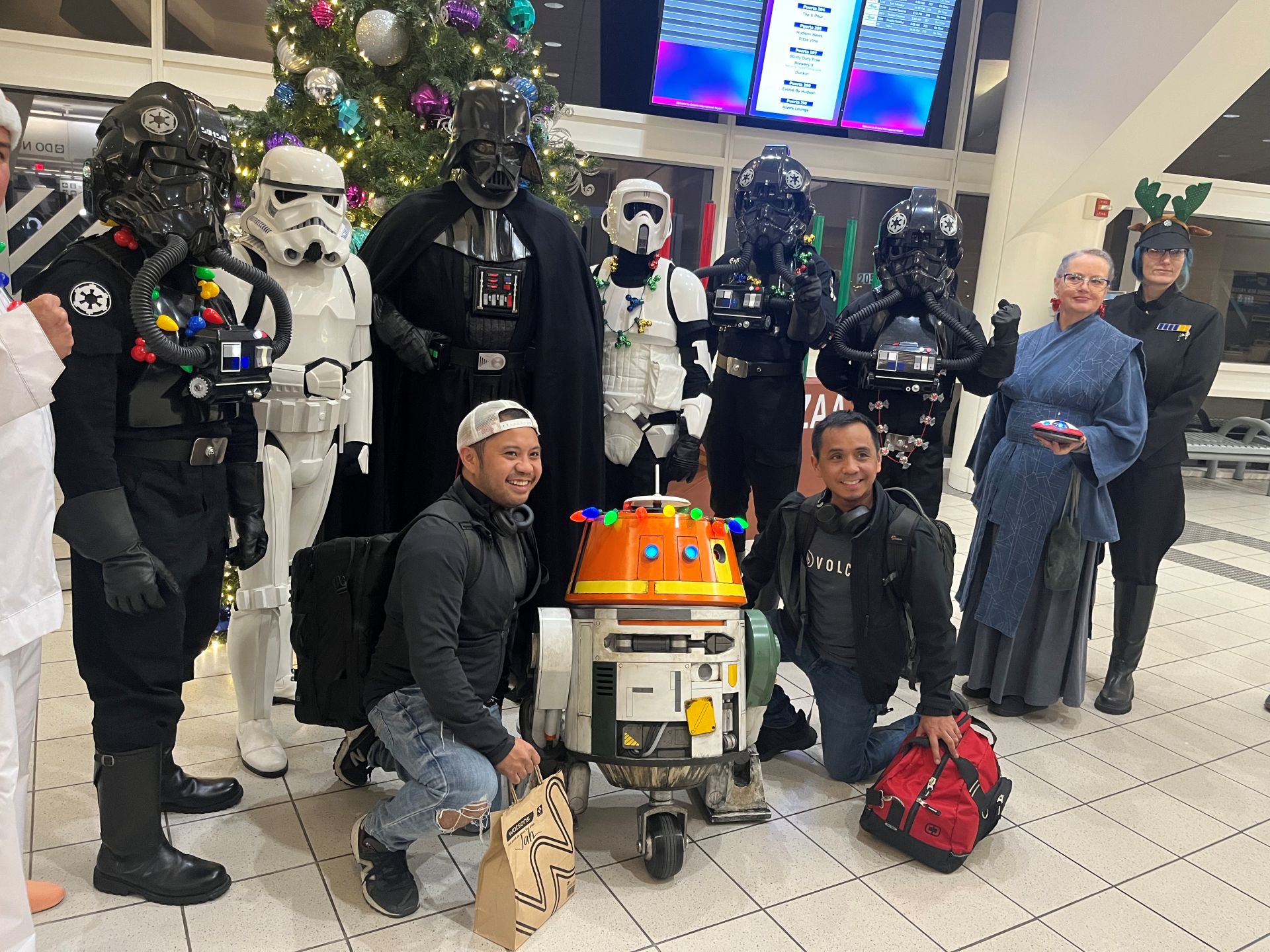 Characters dressed in Star Wars costumes pose with passengers in a terminal with holiday decorations behind them.