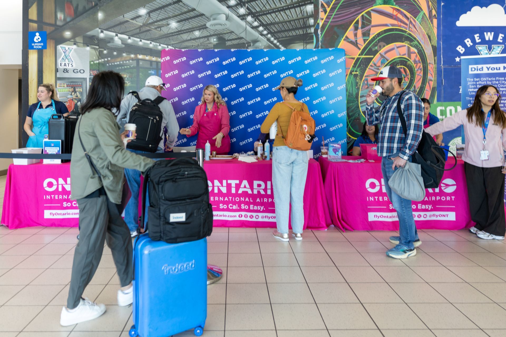 A passenger wheels luggage by tables set up with pink tablecloths that say Ontario International Airport as workers serve coffee to people stopping by.