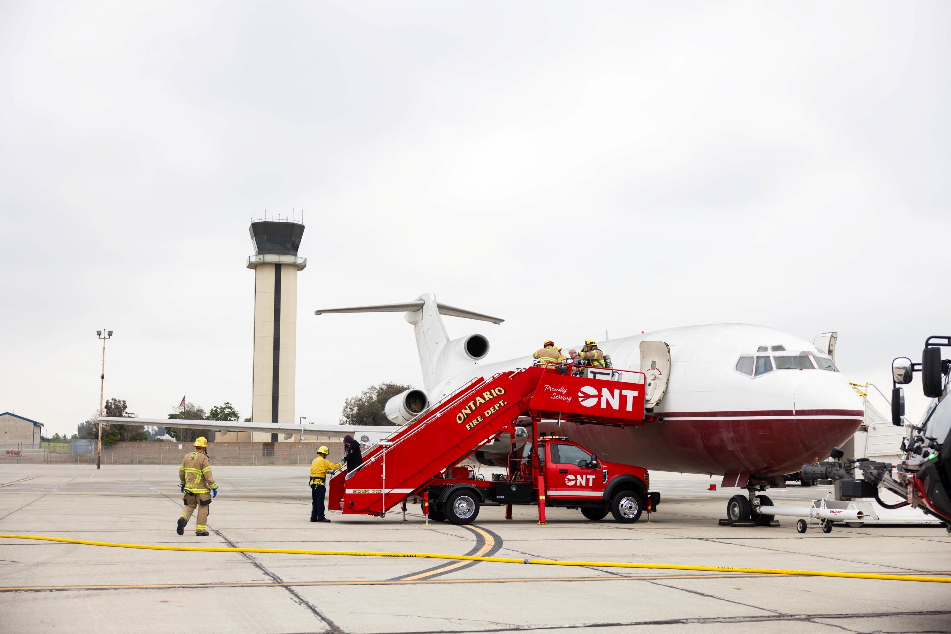 Under cloudy skies, an air traffic tower is seen in the background while a fire department rescue vehicle sits near a plane and firefighters walk nearby.