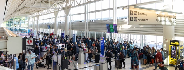 Crowded airport terminal with high ceilings and passengers queuing at check-in counters.