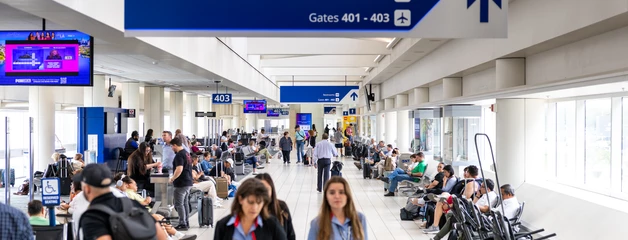 Crowded airport terminal with blue directional signs and passengers waiting at gates.