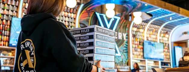 Brewery X employee delivering a stack of pizza boxes across the restaurant with the bar in the background