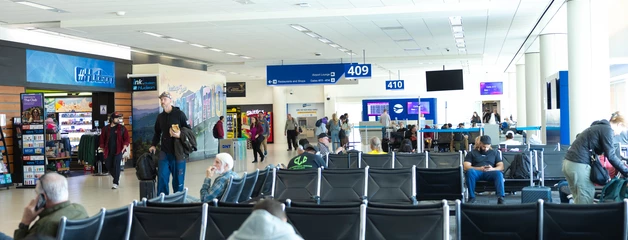 Passengers seated and walking through an airport gate area with visible signs and terminal shops in the background.