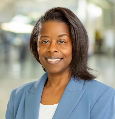 Woman in blue blazer and white top smiling warmly in a bright hallway setting.