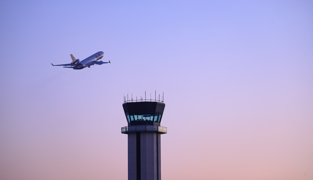 Aircraft flying past airport control tower during sunset with purple sky.