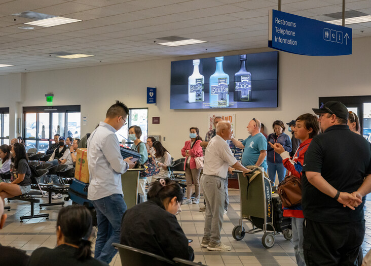 Passengers waiting in line at an airport terminal gate with flight information screens overhead.