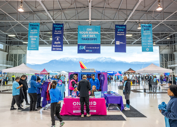 People with luggage standing in an airport terminal with blue directional banners hanging from the ceiling.