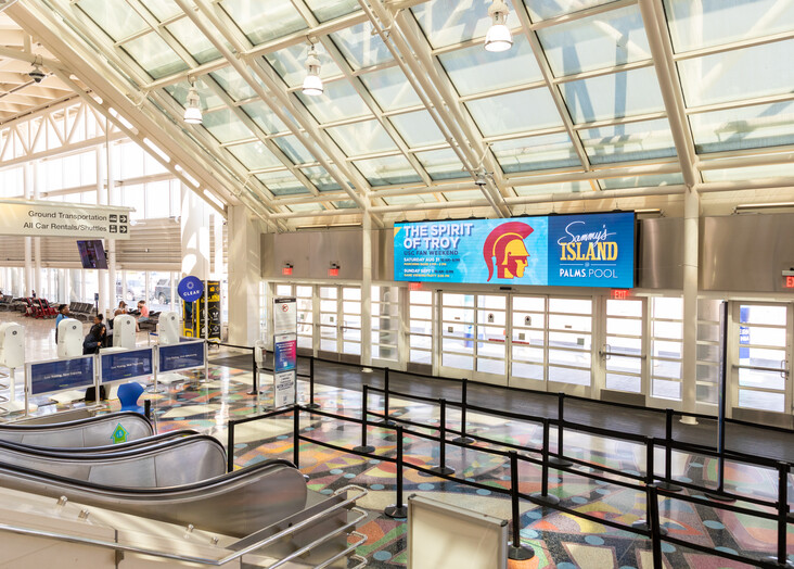 Large, modern airport terminal with glass ceiling, empty security checkpoint area and digital display screens.