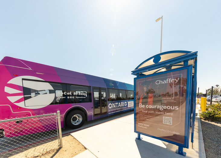 A purple and blue city bus parked at a covered bus stop on a sunny day.